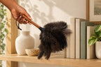 Essential Handheld ostrich feather duster used around books and ceramic objects on a light wood shelf.
