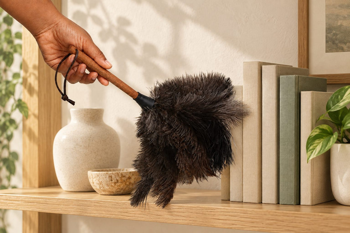 Essential Handheld ostrich feather duster used around books and ceramic objects on a light wood shelf.